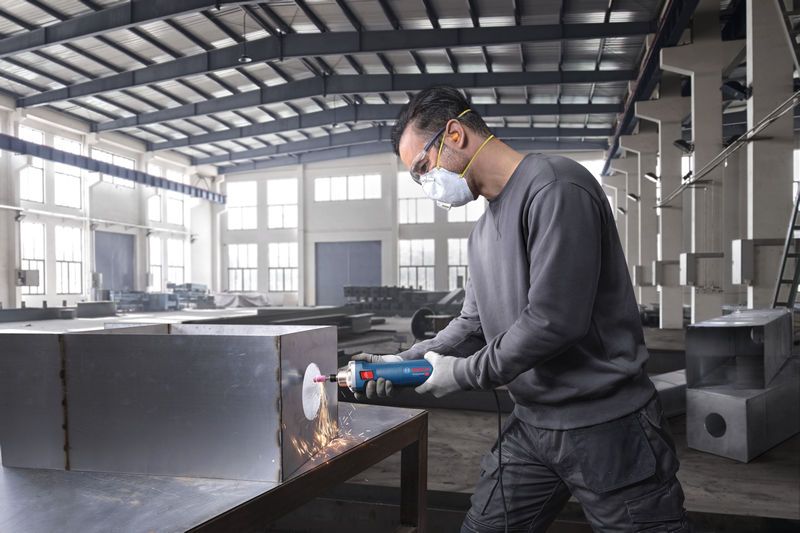 Orang yang mengenakan perlengkapan keselamatan menggunakan penggiling lurus untuk menggiling kotak logam.