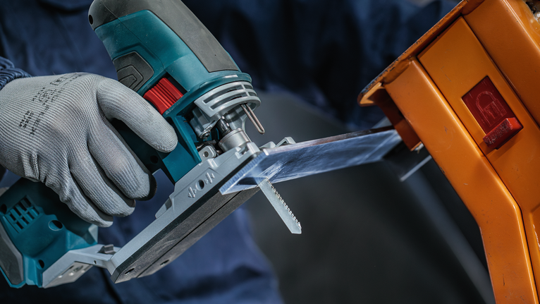 Seseorang yang mengenakan perlengkapan keselamatan menggunakan gergaji ukir untuk memotong lembaran plastik bening.