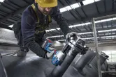 Seseorang yang mengenakan perlengkapan keselamatan menggunakan kunci pas benturan tanpa kabel pada pipa industri.
