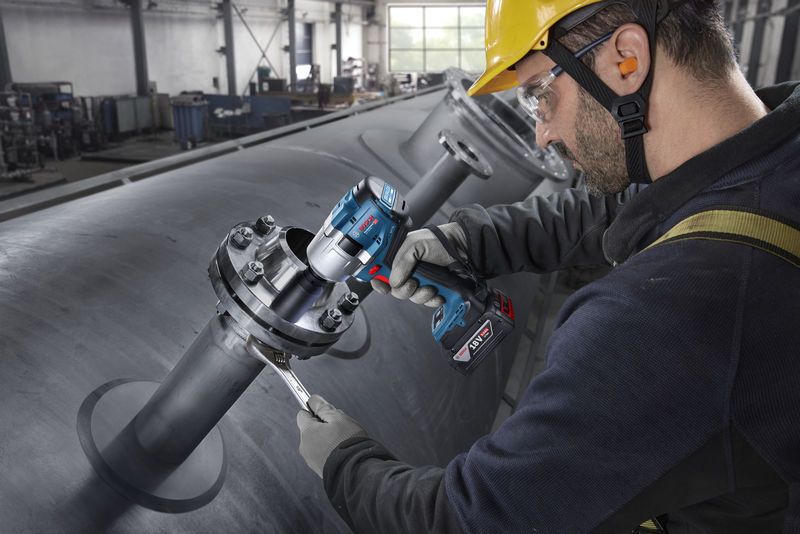 Seseorang yang mengenakan peralatan keselamatan mengencangkan baut pada pipa industri dengan kunci pas benturan tanpa kabel.