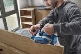 Seseorang yang mengenakan perlengkapan keselamatan menghaluskan papan kayu menggunakan planer listrik.
