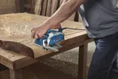 Seseorang menggunakan planer listrik untuk menghaluskan papan kayu di meja kerja.