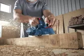 Seseorang yang mengenakan peralatan keselamatan menerbangkan balok kayu, serutan kayu beterbangan.