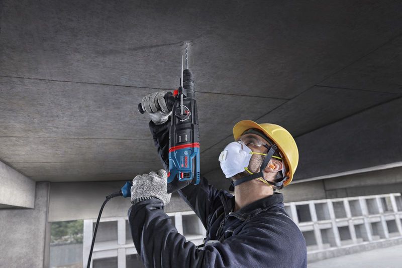 Seseorang yang mengenakan peralatan keselamatan mengebor langit-langit beton menggunakan palu putar.