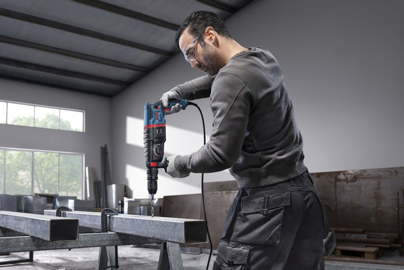 Seseorang yang mengenakan perlengkapan keselamatan mengebor balok baja dengan palu putar.