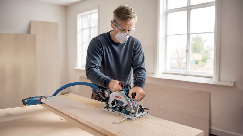 Orang yang mengenakan peralatan keselamatan memotong kayu dengan gergaji bundar tanpa kabel di meja kerja.