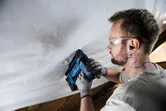 Orang yang mengenakan peralatan keselamatan menggunakan alat tanpa kabel untuk mengencangkan material ke langit-langit.