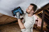Seseorang yang mengenakan perlengkapan keselamatan mengencangkan material ke langit-langit menggunakan alat tanpa kabel.