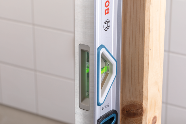 Alat pengukur kerataan (spirit level) memeriksa keselarasan vertikal balok kayu terhadap dinding keramik.