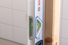 Alat pengukur kerataan (spirit level) memeriksa keselarasan vertikal balok kayu terhadap dinding keramik.
