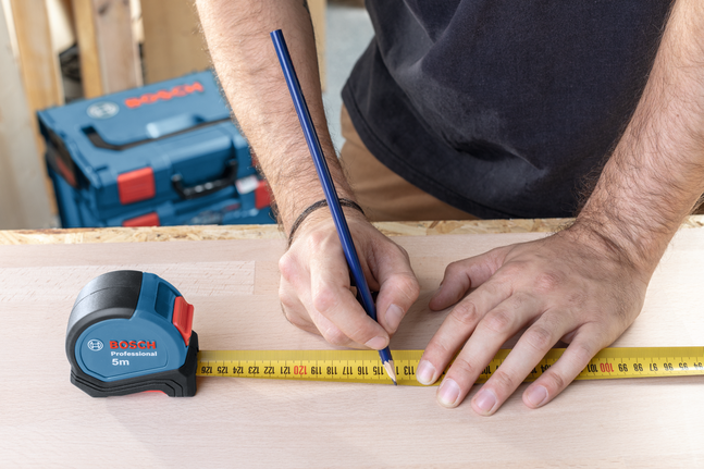 Seseorang sedang menandai ukuran pada kayu menggunakan pita ukur dan pensil.