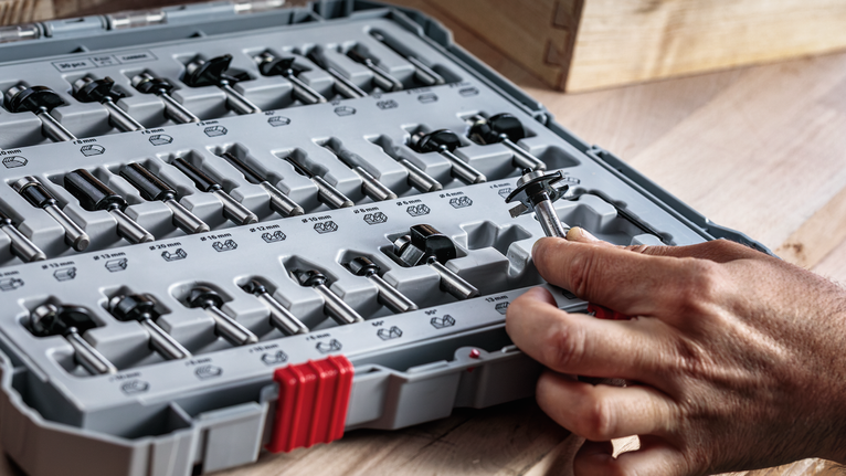Seseorang memilih mata router dari kotak peralatan yang tertata rapi di permukaan kayu.