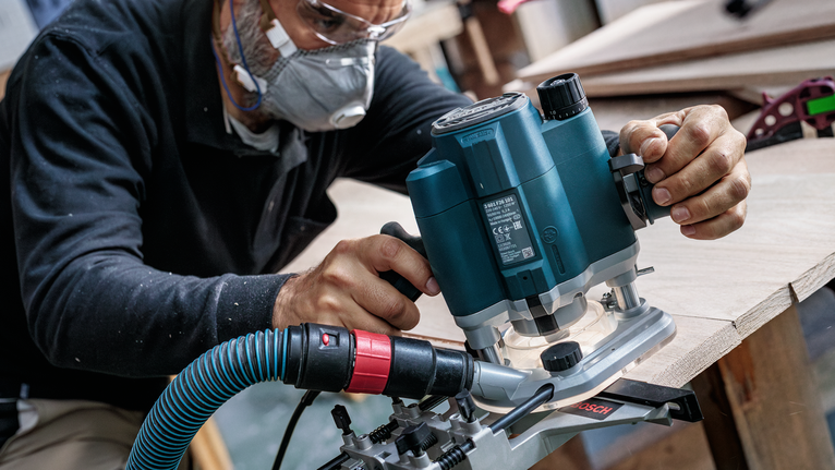 Orang yang mengenakan perlengkapan keselamatan menggunakan router listrik untuk membentuk tepi papan kayu.