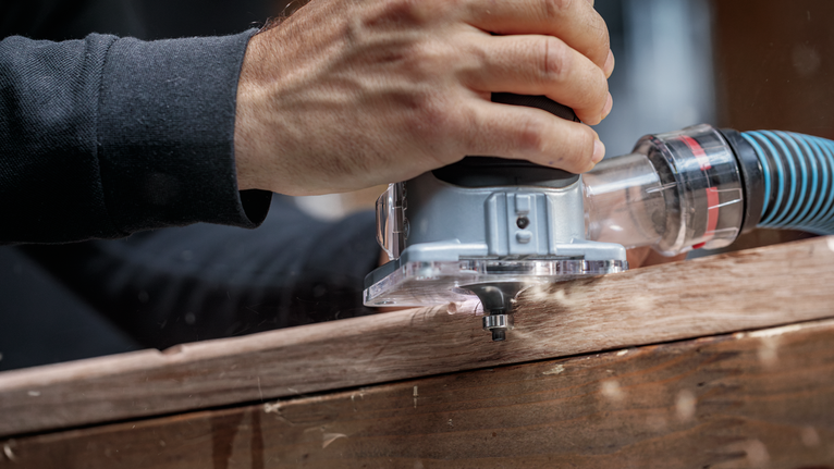 Seseorang memandu router listrik di sepanjang papan kayu untuk membentuk tepi yang presisi.