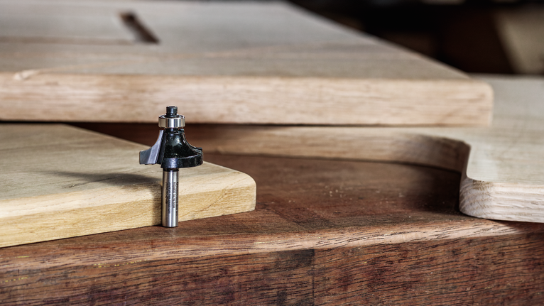 Mata router diletakkan di meja kerja kayu di samping panel kayu yang dipotong dan dibentuk.