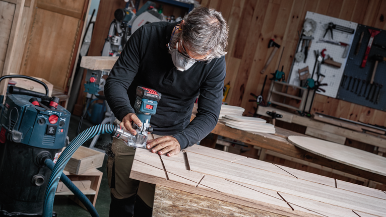 Orang yang mengenakan perlengkapan keselamatan menggunakan alat listrik untuk membuat alur pada papan kayu.