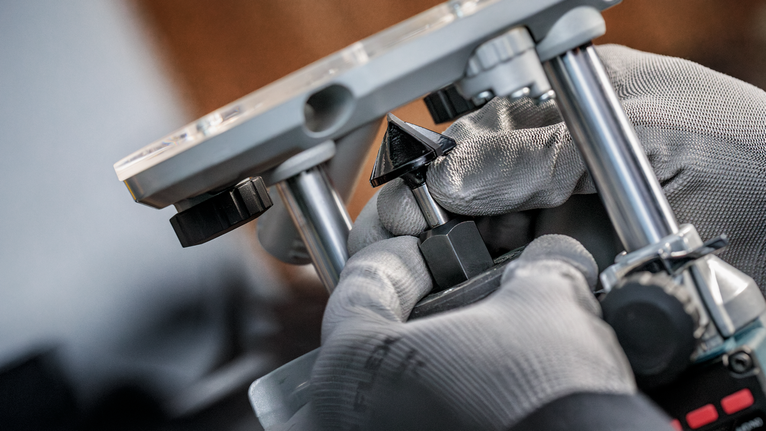 Orang yang mengenakan perlengkapan keselamatan sedang menyetel mata router pada perkakas listrik.