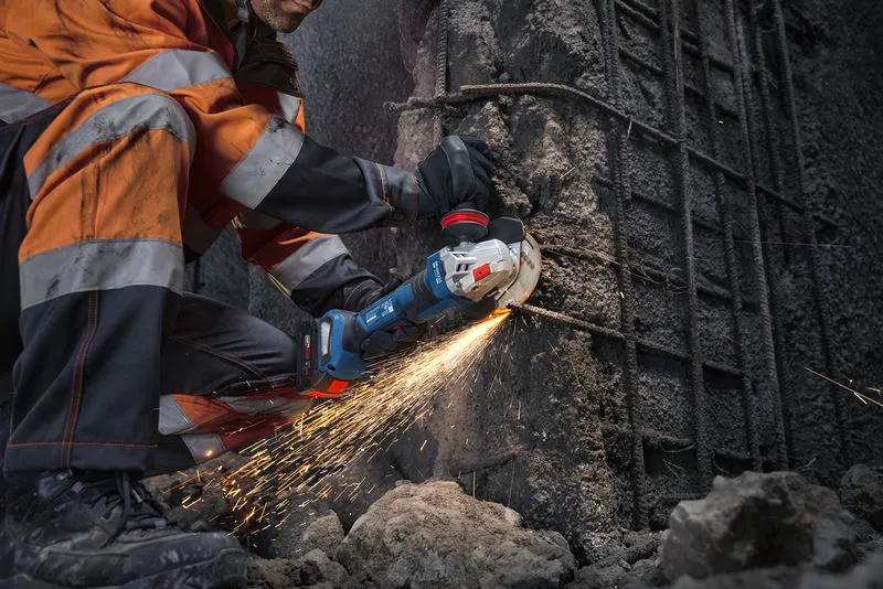 Seseorang yang mengenakan peralatan keselamatan memotong beton dengan mesin gerinda sudut, menghasilkan percikan api.