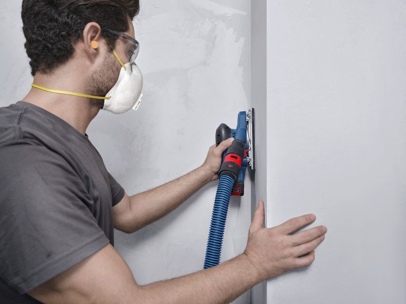 Seseorang yang mengenakan peralatan keselamatan mengampelas dinding dengan mesin pengamplas orbital yang terhubung ke selang vakum.