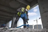 Seseorang yang mengenakan peralatan keselamatan menghancurkan beton dengan palu penghancur.