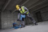 Seseorang yang mengenakan peralatan keselamatan merobohkan lantai beton menggunakan palu penghancur.