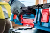 Seseorang yang mengenakan perlengkapan keselamatan mengangkat sistem koper pembawa berwarna biru dan merah.