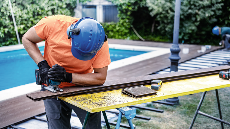 Orang yang mengenakan peralatan keselamatan menggunakan gergaji ukir untuk memotong papan dek di luar ruangan.