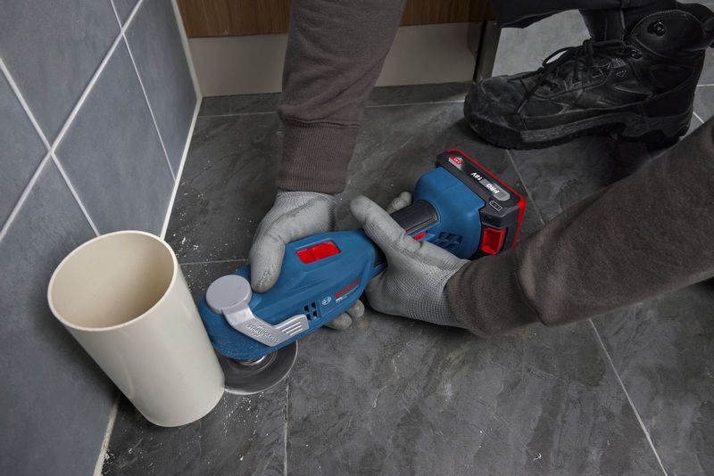 Seseorang yang mengenakan peralatan keselamatan menggunakan alat multifungsi untuk memotong ubin lantai di samping pipa.