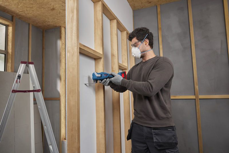 Seseorang yang mengenakan perlengkapan keselamatan menggunakan alat multifungsi untuk memotong drywall.
