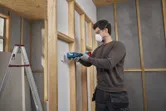 Seseorang yang mengenakan perlengkapan keselamatan menggunakan alat multifungsi untuk memotong drywall.