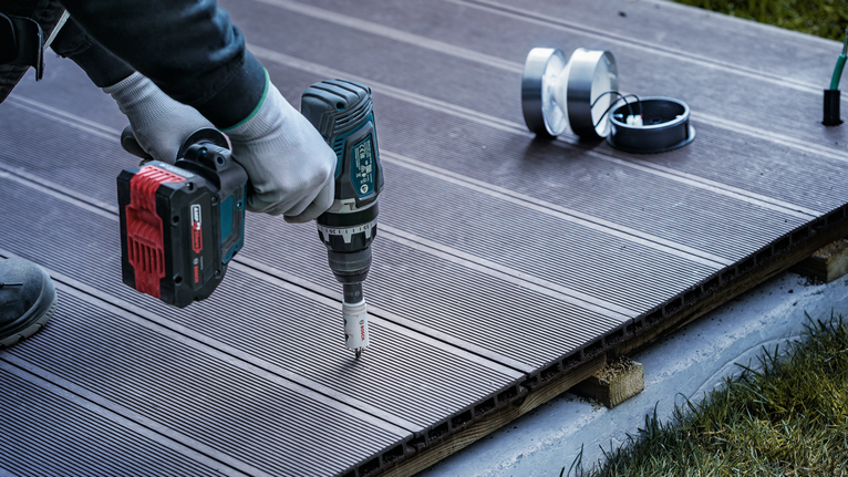 Seseorang yang mengenakan perlengkapan keselamatan menggunakan bor tanpa kabel untuk membuat lubang di dek.