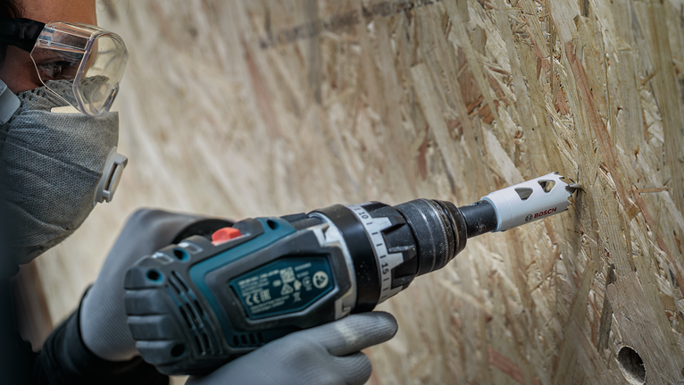 Seseorang yang mengenakan peralatan keselamatan mengebor lubang ke dalam papan partikel.