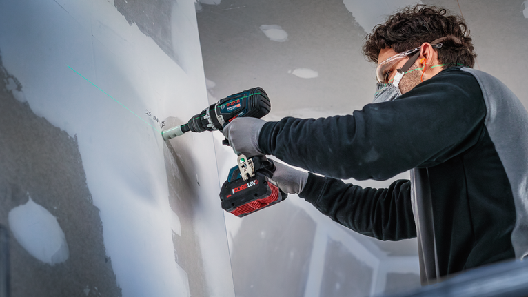 Seseorang yang mengenakan peralatan keselamatan mengebor lubang di dinding gipsum.
