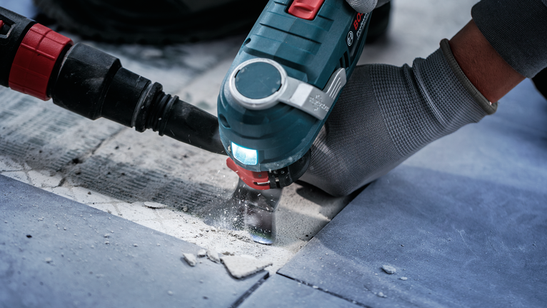 Seseorang yang mengenakan peralatan keselamatan menggunakan perkakas listrik untuk memotong ubin.