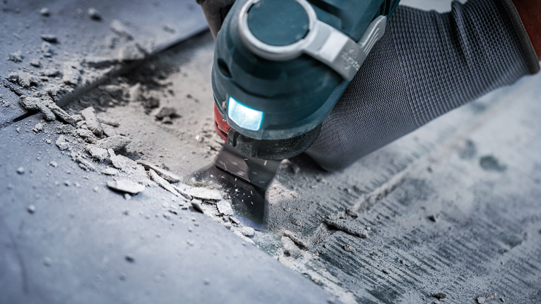 Seseorang yang mengenakan peralatan keselamatan menggunakan alat multifungsi untuk membersihkan material dari lantai.