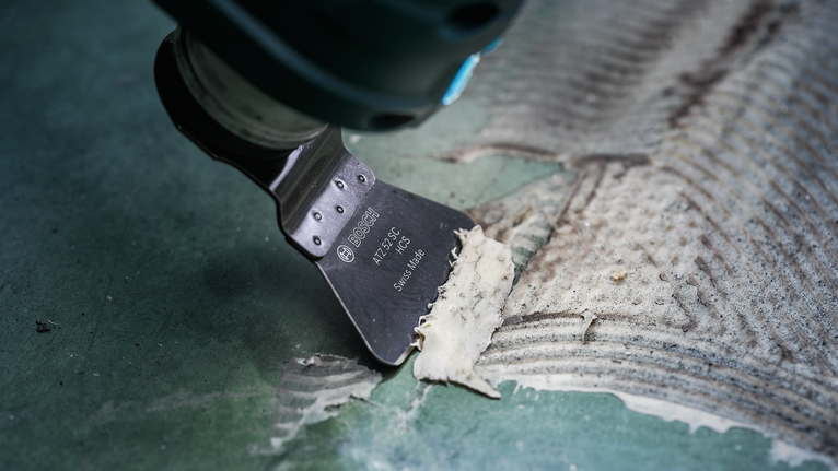 Alat multifungsi berosilasi mengikis sisa perekat dari lantai hijau bertekstur.