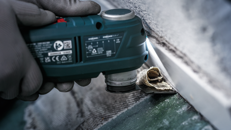 Seseorang yang mengenakan peralatan keselamatan sedang membersihkan lapisan perekat lama dari dinding menggunakan alat multifungsi.