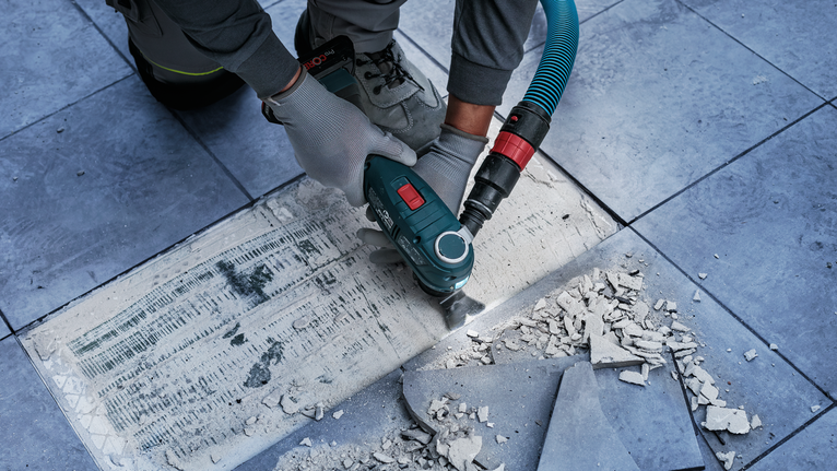 Seseorang yang mengenakan peralatan keselamatan sedang membersihkan perekat ubin dengan alat listrik di lantai keramik.