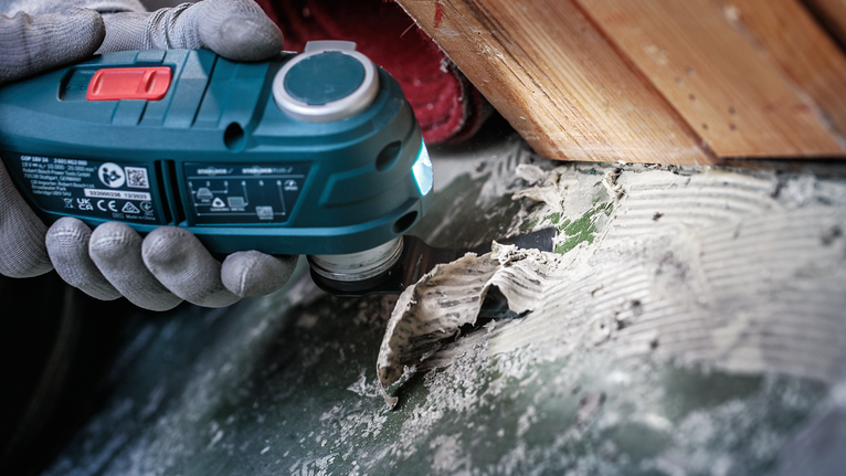 Seseorang yang mengenakan peralatan keselamatan menggunakan alat listrik untuk menghilangkan perekat dari permukaan yang keras.