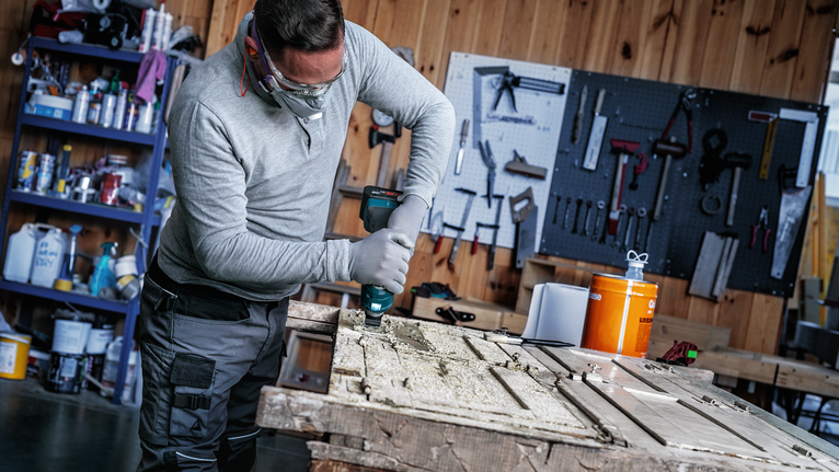 Seseorang yang mengenakan peralatan keselamatan sedang mengebor pintu kayu di atas meja bengkel.