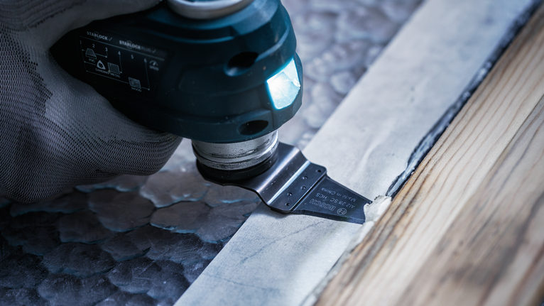 Seseorang yang mengenakan perlengkapan keselamatan menggunakan alat multifungsi untuk memotong material di samping lis kayu.