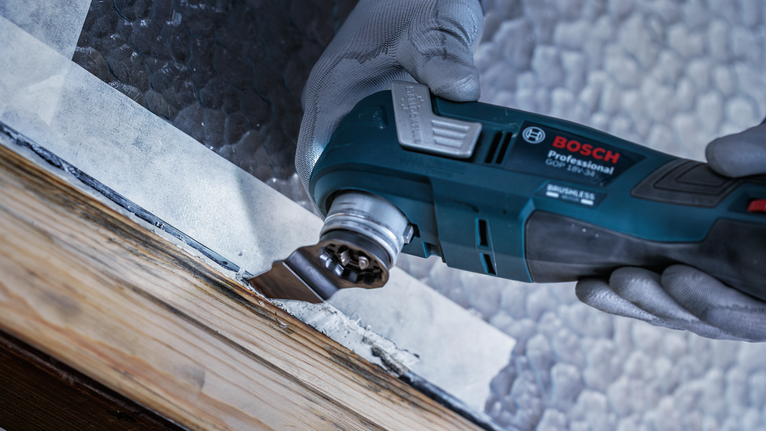 Seseorang yang mengenakan perlengkapan keselamatan menggunakan alat multifungsi untuk mengikis cat dari kusen jendela kayu.