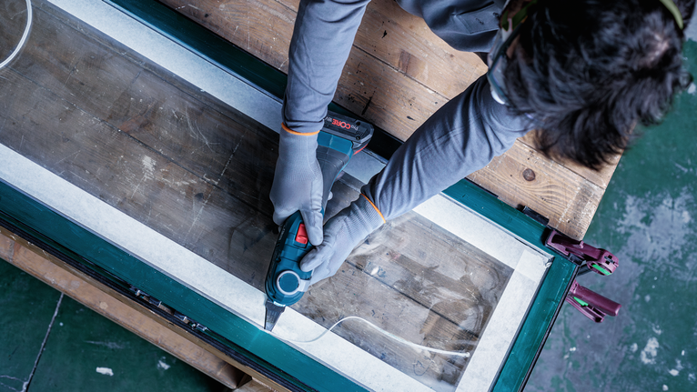 Seseorang yang mengenakan peralatan keselamatan menggunakan perkakas listrik untuk memotong sepanjang bingkai jendela yang telah diberi selotip.