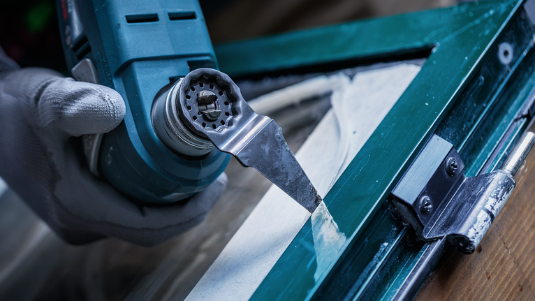 Seseorang yang mengenakan alat pengaman melepas dempul jendela menggunakan alat multifungsi.