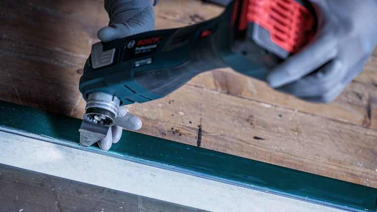 Seseorang yang mengenakan perlengkapan keselamatan menggunakan alat multifungsi untuk mengampelas cat dari kusen jendela.