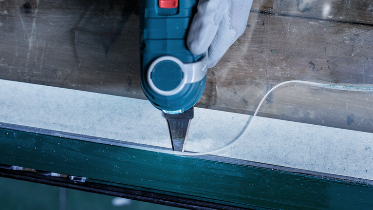 Seseorang yang mengenakan peralatan keselamatan menggunakan alat listrik untuk memotong garis perekat di sepanjang jendela.