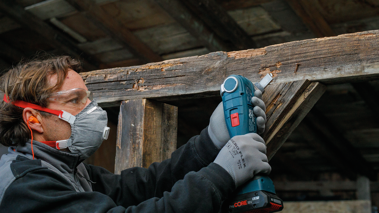 Orang yang mengenakan peralatan keselamatan menggunakan alat tanpa kabel untuk mengampelas balok kayu.