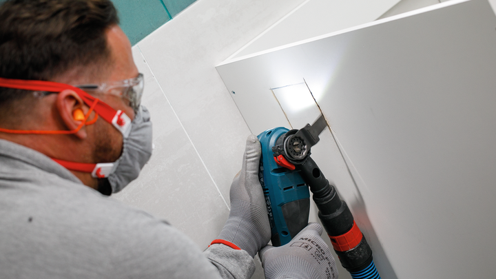 Orang yang mengenakan peralatan keselamatan memotong lubang persegi panjang pada panel dengan alat listrik.