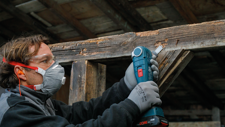 Orang yang mengenakan peralatan keselamatan menggunakan alat listrik untuk mengampelas balok kayu.