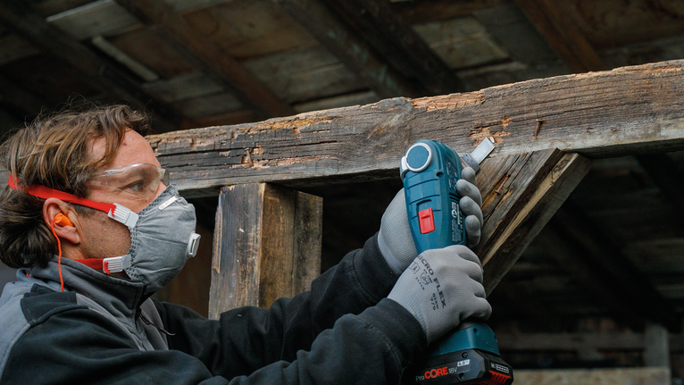 Orang yang mengenakan peralatan keselamatan menggunakan alat listrik untuk mengampelas balok kayu.
